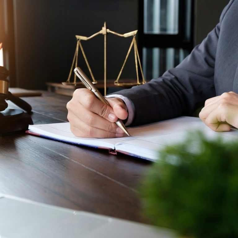Person in Anzug schreibt in ein Notizbuch, im Hintergrund eine Waage als Symbol für Recht und Gerechtigkeit.